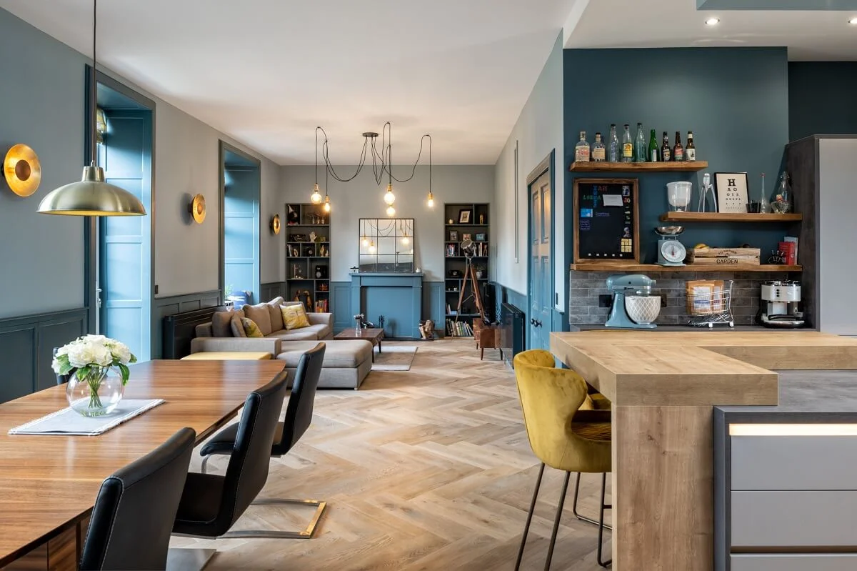 Russwood European Oak Flooring in Dune, laid in a herringbone arrangement in a large open plan living and kitchen area with a sofa, and colourful blue walls.