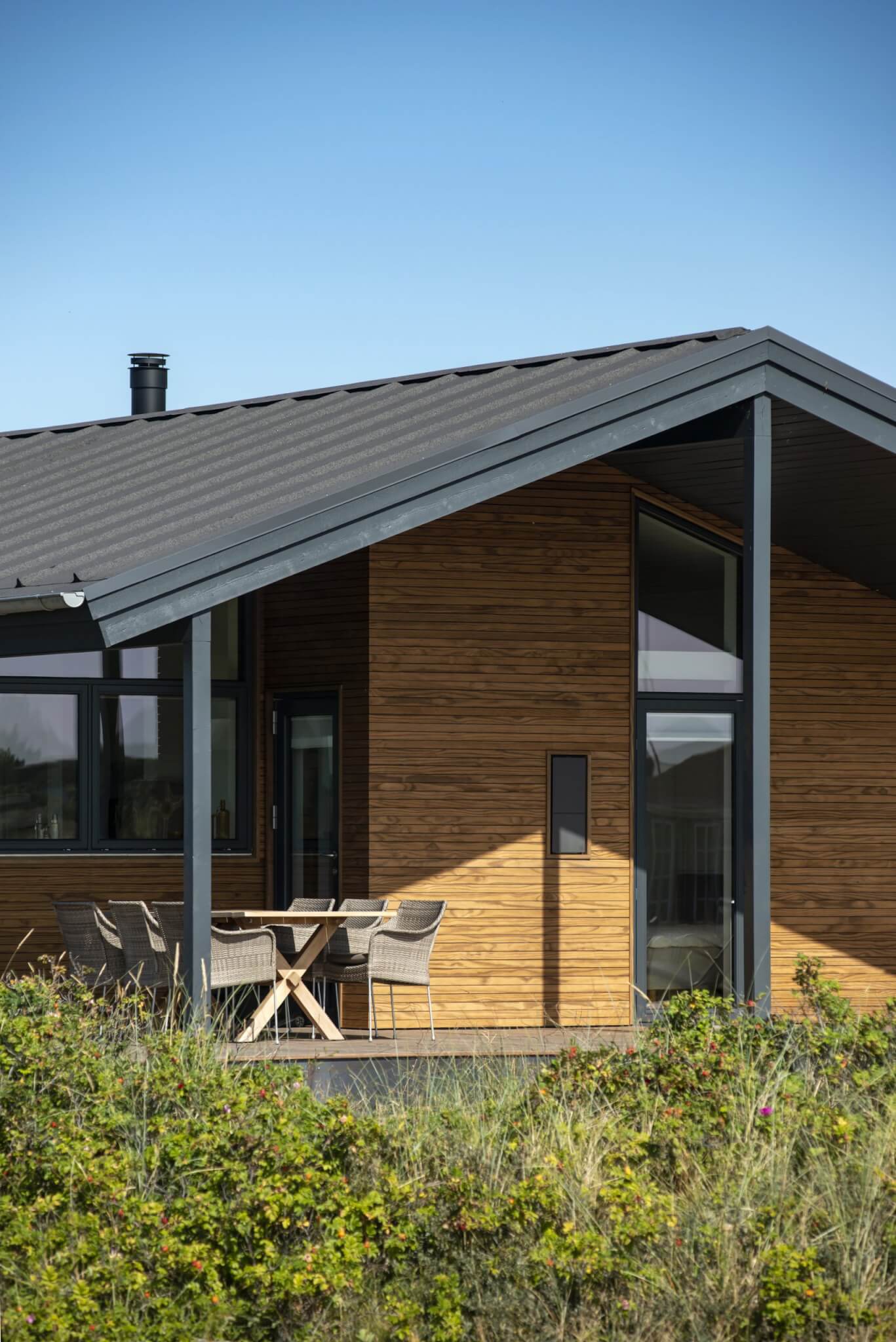 A timber clad house amongst greenery with outdoor chairs.